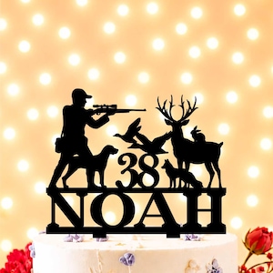 May include: Black cake topper with a hunter, dog, ducks, deer, fox, and rabbit silhouettes. The number "38" and the name "NOAH" are displayed. The topper is on a decorated cake, suitable for a birthday celebration.