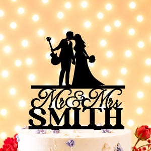 May include: A black cake topper featuring a silhouette of a couple kissing, with one person holding a guitar. Below the figures, the text reads Mr & Mrs SMITH in a combination of cursive and block lettering, all mounted on a horizontal base.