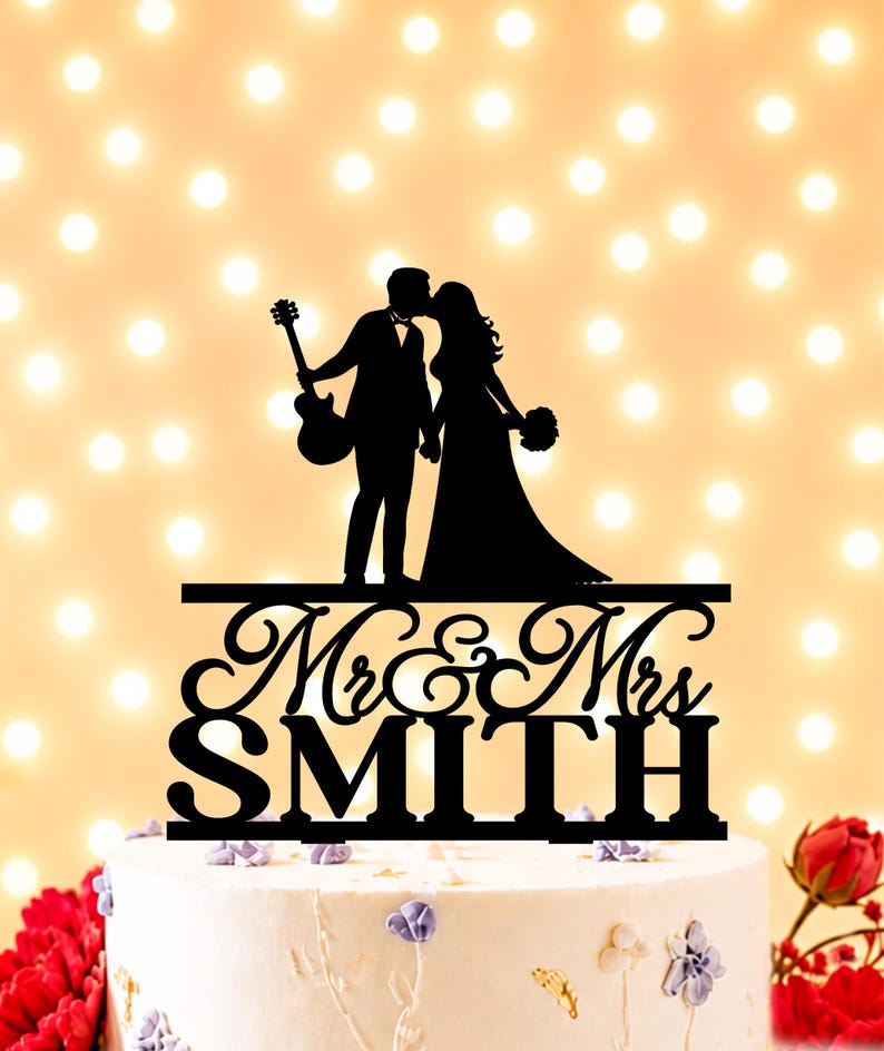 May include: A black cake topper featuring a silhouette of a couple kissing, with one person holding a guitar. Below the figures, the text reads Mr & Mrs SMITH in a combination of cursive and block lettering, all mounted on a horizontal base.