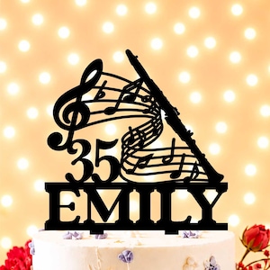Peut inclure: Décoration de gâteau noire sur le thème de la musique. Elle comprend une clé de sol, des notes de musique, une flûte, le chiffre 35 et le nom "EMILY". La décoration est placée sur un gâteau décoré, avec un arrière-plan flou.