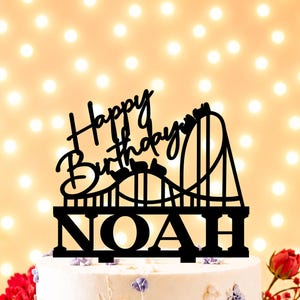 Puede incluir: Adorno negro para pastel con las palabras "Happy Birthday" y "NOAH" en fuente cursiva. El adorno presenta un diseño de montaña rusa. El pastel está decorado con flores moradas y hojuelas doradas.