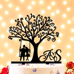 Könnte beinhalten: Ein schwarzer Tortenaufsatz mit der Silhouette von zwei Figuren auf einer Schaukel, die an einem Baum mit herzförmigen Blättern hängt. Rechts neben dem Baum sind die Buchstaben A und S durch ein Herz verbunden, alles auf einer flachen Basis.