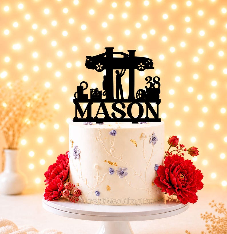Peut inclure: Un g&acirc;teau blanc orn&eacute; de d&eacute;corations florales et surmont&eacute; d'un cake topper noir. Le cake topper repr&eacute;sente une voiture sur un pont &eacute;l&eacute;vateur, une personne et le nom "MASON" avec le chiffre "38". Des fleurs rouges et un pr&eacute;sentoir &agrave; g&acirc;teau blanc compl&egrave;tent le design.