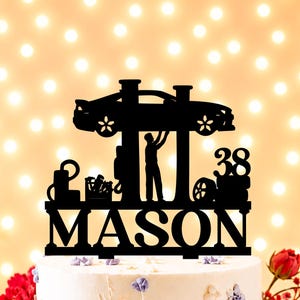 Peut inclure: D&eacute;coration de g&acirc;teau noire repr&eacute;sentant une voiture en r&eacute;paration par un m&eacute;canicien, le nom "MASON" et le chiffre "38". Le g&acirc;teau est blanc, orn&eacute; de fleurs violettes et dor&eacute;es et de roses rouges.
