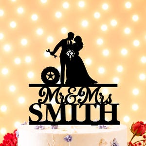 Könnte beinhalten: Ein schwarzer Tortenaufsatz als Silhouette, der ein stehendes Paar zeigt, wobei eine Person einen Schraubenschlüssel hält und ein großer Reifen daneben steht. Unter den Figuren steht der Text Mr & Mrs SMITH in Schreib- und Blockschrift.