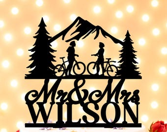 May include: A black cake topper featuring a silhouette of two people standing with bicycles between two pine trees and a mountain range. Below the scene, the text reads Mr & Mrs WILSON in a combination of cursive and bold capital lettering.