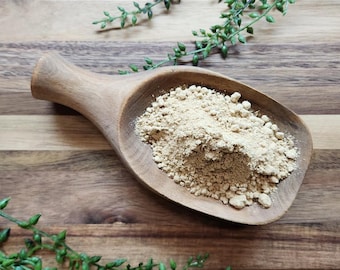 Puede incluir: Una pala de madera de color marrón claro llena con un montón de polvo fino de color beige. La pala tiene un mango redondeado y liso, y un cuenco profundo de forma ovalada que sostiene la sustancia seca y granular en su textura.