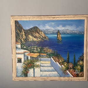 Könnte beinhalten: Ölgemälde einer Küstenlandschaft mit blauem Meer, Bergen und weißen Gebäuden. Das Kunstwerk ist in einem verwitterten, cremefarbenen Holzrahmen gerahmt. Das Gemälde zeigt einen sonnigen Tag mit klarem Himmel und einer kleinen Insel im Meer.