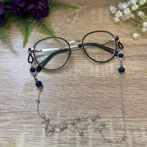 May include: A pair of round-framed spectacles with a silver-toned metal chain attached to the temples. The chain features small silver beads and dark blue spherical beads, connecting to the glasses with black rubber loops on each side.