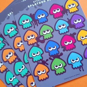 Puede incluir: Un conjunto de 16 pegatinas coloridas con personajes de calamar de dibujos animados del videojuego Splatoon. Las pegatinas están impresas sobre un fondo gris con tinta negra. Las pegatinas están dispuestas en una cuadrícula y presentan una variedad de colores, incluidos el rojo, el azul, el verde, el naranja y el morado.