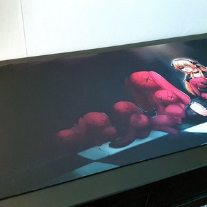 Op de afbeelding: Een zwart-witte muismat met een donkere achtergrond waarop een jonge vrouw met lang rood haar te zien is die een zwarte jurk en een koptelefoon draagt. Ze wordt omringd door een groep roze pluche speelgoed met X'en voor ogen.