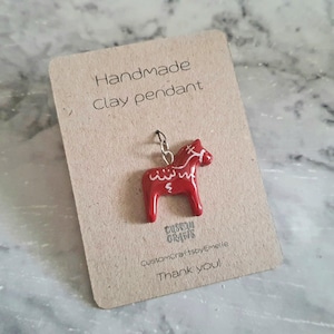 Könnte beinhalten: Handgefertigter Tonanhänger mit einem roten Dala-Pferd mit weißen Akzenten. Der Anhänger ist an einer silberfarbenen Kette befestigt und wird auf einer braunen Karte mit dem Text "Handmade Clay pendant" präsentiert.