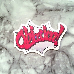 May include: A white sticker with a red exclamation point and the word "Objection!" in a bold, stylized font. The sticker has a sparkly, glittery effect.
