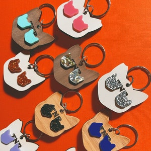 May include: A collection of cat-shaped keychains in various colors and materials. The keychains feature a cat face design with two cat head shapes as accents. Colors include pink, blue, silver glitter, and brown wood. Each keychain has a silver ring.