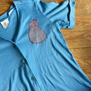 May include: A light blue button-up shirt with a gingham pear patch on the front.