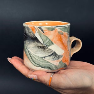 May include: A ceramic mug with a marbled design in shades of black, green, and orange. The mug has a rounded shape and a handle.