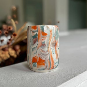 Peut inclure: Un petit vase en céramique blanche avec un motif marbré dans des tons d'orange, de brun et de vert. Le vase a une forme légèrement irrégulière.