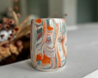 Op de afbeelding: Een kleine, witte keramische vaas met een marmerpatroon in tinten oranje, bruin en groen. De vaas heeft een licht onregelmatige vorm.