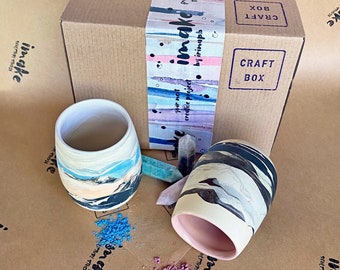 Könnte beinhalten: Zwei Keramikbecher mit abstrakten Marmor-Designs in Blautönen, Weiß und Schwarz. Die Becher stehen auf einer braunen Papieroberfläche mit rosa und blauem Pulver. Ein brauner Karton mit dem Text "CRAFT BOX" und "imake" befindet sich im Hintergrund.
