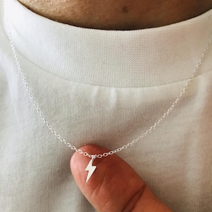 Puede incluir: Un collar de plata con un colgante de rayo. El colgante cuelga de una cadena fina. El collar se muestra sobre una camiseta blanca.