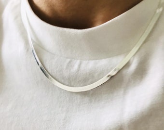 Puede incluir: Un collar de cadena de plata que se lleva alrededor del cuello de una persona. La cadena es fina y tiene una forma rectangular plana.
