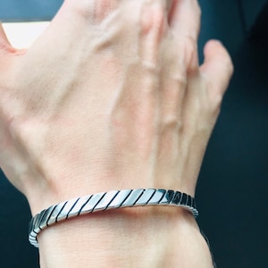 May include: A silver bracelet with a textured, diagonal pattern. The bracelet is open and has a simple, minimalist design.