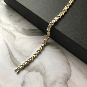 Puede incluir: Un brazalete de tono plateado y dorado con un diseño de cadena de eslabones. El brazalete está sobre una superficie de mármol.