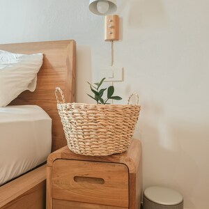 Può includere: Un cesto di vimini intrecciato con una piccola pianta verde all'interno si trova su un comodino di legno accanto a un letto con biancheria da letto bianca. Il comodino ha due cassetti e un piano in legno.