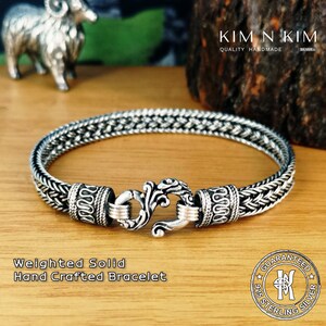May include: A silver bracelet with a braided chain design and a decorative clasp. The bracelet is labeled "Weighted Solid Hand Crafted Bracelet" and "925 Sterling Silver Guaranteed".