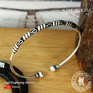 以下が含まれることがあります： 複雑なディテールが施されたシルバーのバリ・ジャワ・トルクバングルブレスレット。ブレスレットはオープンタイプで、丸みを帯びた端になっています。画像の右上隅には「KIM N KIM QUALITY HANDMADE SILVER」というテキストが表示されています。画像の右下隅には「GUARANTEED 925 STERLING SILVER」というテキストが表示されています。