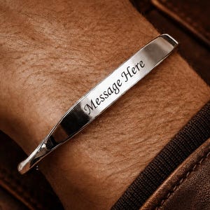 Puede incluir: Una pulsera tipo brazalete de metal plateado pulido con una superficie lisa y plana que presenta el texto "Message Here" grabado en una fuente cursiva oscura y elegante. La pulsera tiene un diseño sencillo y abierto sobre la piel.