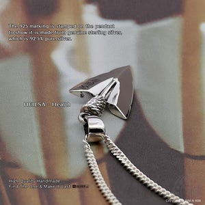May include: A silver arrowhead pendant on a chain. The pendant features a textured rope-like detail at the top and a smooth, polished surface. The text HEILSA - Health is visible, along with a 925 stamp indicating 92.5% pure sterling silver.