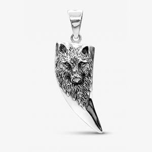 May include: A silver-toned pendant shaped like a curved tooth or claw, featuring a detailed, raised relief of a wolf's head on the upper portion. The metal has a polished finish with darkened recessed areas to highlight the texture of the fur.