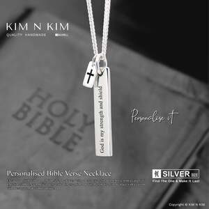 May include: A silver necklace featuring a rectangular pendant engraved with "God is my strength and shield." A small cross charm is attached. The necklace is shown against a blurred background.
