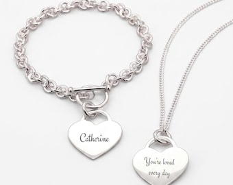 Puede incluir: Una pulsera de eslabones plateados con cierre de barra y un colgante en forma de corazón grabado con el nombre Catherine, junto a un collar de cadena plateada con un colgante en forma de corazón grabado con el texto You're loved every day.