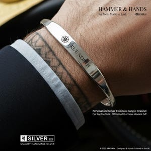 Könnte beinhalten: Ein silbernes Armreif-Armband mit der Gravur "TRUE NORTH" und einer Kompassrose. Das Armband befindet sich an einem Handgelenk. Der Text "HAMMER & HANDS" befindet sich über dem Armband. Unisex-Armreif.