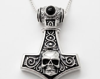 May include: A silver-toned metal pendant shaped like a hammer, featuring an embossed skull at the center, intricate scrollwork patterns, and a smooth black circular stone set at the top. The pendant hangs from a matching silver-toned metal chain.