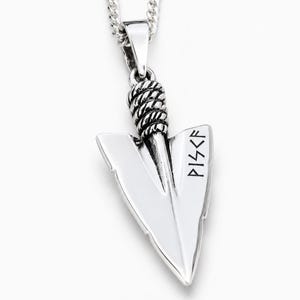 May include: A silver-toned metal pendant shaped like an arrowhead, suspended from a matching chain. The pendant features a twisted rope-like texture at the top and the letters PISCF engraved in a dark, angular font on the right side of the blade.