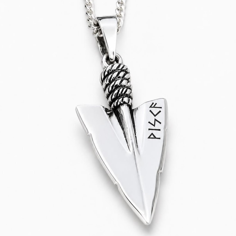 May include: A silver-toned metal pendant shaped like an arrowhead, suspended from a matching chain. The pendant features a twisted rope-like texture at the top and the letters PISCF engraved in a dark, angular font on the right side of the blade.