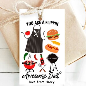 Puede incluir: Una tarjeta blanca con una ilustración en blanco y negro de una parrilla, una hamburguesa, un perrito caliente, un tomate y un chile. El texto en la tarjeta dice "You are a Flippin' Awesome Dad love from Harry".