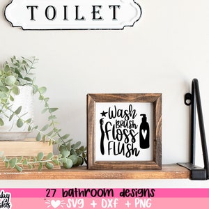 May include: A wooden sign with the text "Wash Brush Floss Flush" in black lettering. The sign is on a shelf with a plant and a sign that says "Toilet".