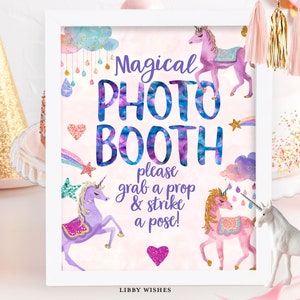 May include: A white framed sign with a pink background and the text "Magical Photo Booth" in blue and purple. The sign also includes two unicorns, one purple and one pink, with rainbow accents and the text "Please grab a prop & strike a pose!" in purple.