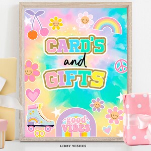 以下が含まれることがあります： タイダイの背景に「CARD'S and GIFTS」の文字が大きくカラフルに書かれた額入りのサイン。花、虹、さくらんぼ、ローラースケート、「good vibes」の文字で飾られています。