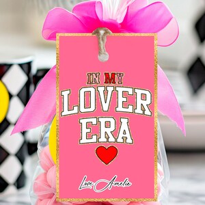 Può includere: Etichetta regalo rosa con bordo dorato glitterato e il testo "In My Lover Era" in lettere bianche con un cuore rosso. L'etichetta è legata con un nastro rosa e ha il testo "Love, Amelie" in corsivo.