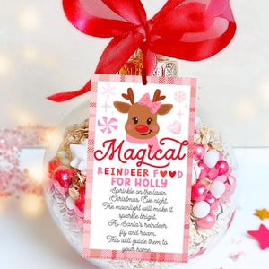 May include: A clear glass ornament filled with decorative elements, topped with a red bow. A pink and white tag reads "Magical Reindeer Food for Holly" with a reindeer graphic. The ornament contains pink and white beads.