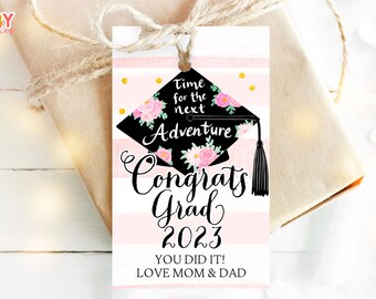 May include: Pink and white striped graduation gift tag with a black graduation cap decorated with pink flowers. The tag says "Time for the next Adventure Congrats Grad 2023 You Did It! Love Mom & Dad".