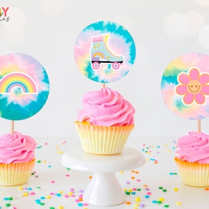 Puede incluir: Tres cupcakes con glaseado rosa y envoltorios amarillos. Cada cupcake tiene una decoración diferente con temática tie-dye: un arcoíris, un patín y una flor con una cara sonriente.