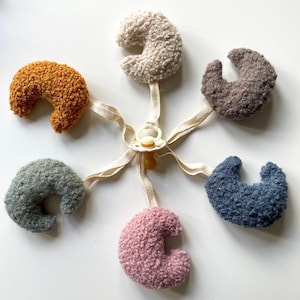 Puede incluir: Un juguete para bebé con seis piezas de felpa en forma de media luna de diferentes colores, unidas a un clip para chupete. Los colores son beige, naranja, marrón, gris, azul y rosa.