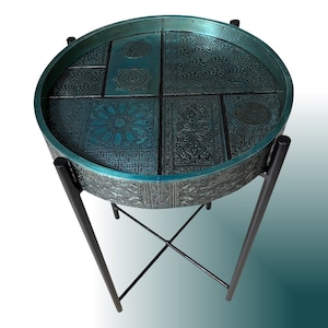 Pode incluir: Uma mesa auxiliar redonda azul turquesa com um design em relevo intrincado. A mesa tem uma estrutura de metal preto com um design em forma de cruz.
