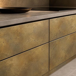 Pode incluir: Armários de cozinha com portas de painel plano e acabamento em latão metálico envelhecido. Os armários estão dispostos numa fila horizontal sob uma bancada de pedra cinzenta, que contém uma taça escura e uma placa de indução preta.